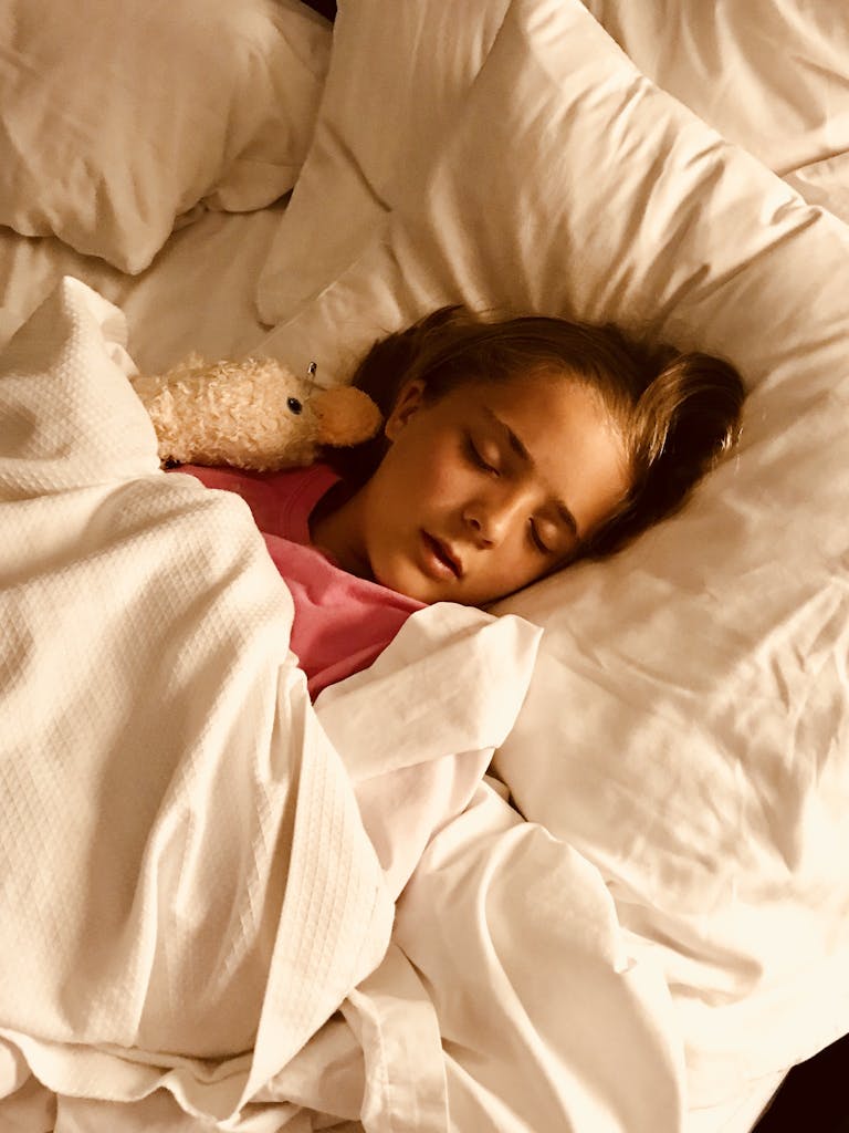 Young girl sleeping peacefully with stuffed animal in a cozy white bed.